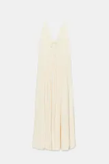 Vestido largo satinado color crudo, con escote en pico adornado con lazada y tirantes finos ajustables. Presenta detalles de frunces y diseño fluido.