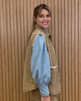 Chaleco de denim color marrón, confeccionado en 100% algodón, con bolsillos frontales amplios, cierre de botones y costuras decorativas bordadas en blanco en los bordes y los bolsillos.