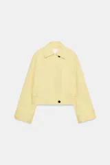 Chaqueta corta confeccionada en mezcla de algodón color amarillo claro, con cuello de solapa, manga larga, bolsillos delanteros y cierre frontal con botones visibles en los extremos.