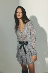 Vestido corto de tejido transparente, con estampado de patrón geométrico y floral en tonos azules y blancos, cuello redondo con pequeña abertura y tiras para atar. Tiene manga larga abullonada, cintura elastizada y falda con volados en capas.