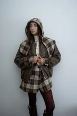 Chaqueta de cuero sintético color marrón con capucha y solapas amplias en tela escocesa a cuadros. Posee cierre frontal, puños elásticos y diseño de corte holgado.