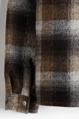 Sobrecamisa acolchada de corte relajado con diseño a cuadros en tonos marrones, grises y negros. Presenta cuello inglés, cierre frontal con botones a presión, hombros caídos y mangas largas con puños abotonados.