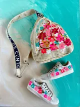 Mochila Kiri de cuero natural de la marca Carolina De Cunto, con diseño artístico pintado a mano de flores multicolores en el frente. Cuenta con una correa bandolera negra con el nombre de la marca en letras blancas y cierre superior con cremallera.