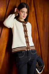 Cardigan de punto color crudo con detalles en marrón y negro. Presenta cuello redondo, cierre frontal con botones forrados y puños acanalados con rayas horizontales.