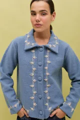 Chaqueta de lana merino hervida color marrón, con cuello camisero y botones forrados. Presenta bordados artesanales en color ocre que recorren el frente, el cuello y los puños.