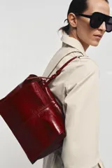 Bolso formato mini shopper de piel con acabado brillante y efecto arrugado. Cuenta con doble compartimento, cierre mediante botón imantado, bolsillo interior y asa de hombro ajustable.