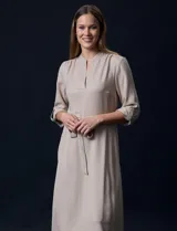 Vestido maxi color beige, con cuello mao y escote en V. Presenta mangas largas con presilla y botón para regular el largo, y un lazo ajustable en la cintura que marca la silueta.