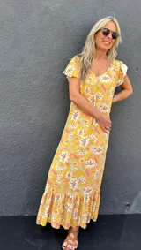 Vestido largo amarillo con estampado floral en tonos rosa, blanco y celeste. Tiene mangas cortas con volados y un volante en el ruedo.