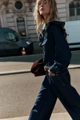 Camisa de denim azul oscuro con cuello redondo y manga larga. Presenta un diseño con volantes en el pecho y hombros, puños con botones y cierre frontal con botones dorados.