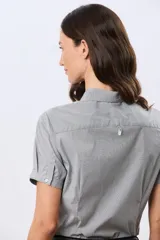 Camisa celeste rayada de manga corta, con cuello camisero y cierre frontal con botones. Presenta un corte entallado y un pequeño detalle de placa metálica en el borde inferior.