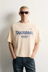 Camiseta relaxed fit de color beige, con cuello redondo, mangas cortas y estampado frontal en tono azul con texto gráfico.