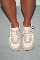 Zapatillas deportivas estilo barefoot con puntera amplia y suela plana flexible. Presentan un diseño combinado en tonos beige y crema, con paneles de gamuza y tela, cierre mediante cordones bicolor y suela con efecto engomado.