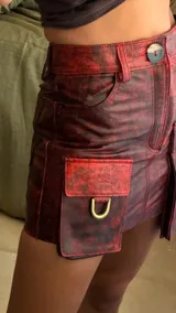 Minifalda de cuero con efecto desgastado en tonos rojos y negros, corte recto y cintura alta con trabillas. Presenta bolsillos cargo laterales con solapa y detalles de argollas metálicas doradas.