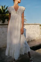 Vestido largo color crudo, sin mangas, con escote en V pronunciado, canesú superior de tejido de punto con botones frontales y lazo para atar en el cuello, y falda fluida con detalle de bordado en el bajo.