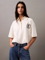 Chomba color crema de punto de algodón con cuello clásico, tapeta de botones oculta y mangas cortas. Presenta un corte holgado y un escudo bordado con el logo de Calvin Klein en el pecho.