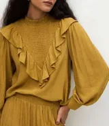 Blusa de manga larga color mostaza con cuello alto y detalle de panel delantero texturizado. Presenta volados aplicados en el busto y puños abotonados.