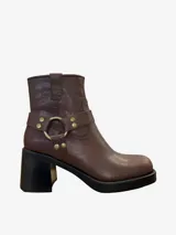 Botín de cuero marrón con tacón de bloque grueso y plataforma. Presenta una tira decorativa con argolla metálica dorada y tachuelas alrededor del tobillo, estilo motociclista.