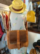 Morral de cuero gamuzado color tostado, con correa regulable, solapa frontal de pelo vacuno y cierre central con tira vertical.