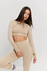 Conjunto deportivo de dos piezas en color beige, compuesto por un top de manga larga con aberturas para pulgares y calzas de tiro alto. Ambas prendas presentan un diseño sin costuras con detalles texturizados y el logo de la marca estampado en el frente del top.