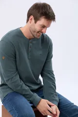 Remera de algodón texturada color gris oscuro, de manga larga, con cuello redondo y botones en la parte frontal. Presenta un pequeño logo bordado en el pecho y un parche decorativo en la manga izquierda.