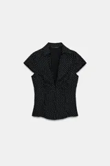 Camisa entallada de popelín negro con estampado de lunares blancos, escote en pico, cuello solapa y mangas muy cortas. Cuenta con costuras marcadas y cierre lateral con cremallera oculta.
