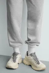 Zapatilla deportiva tipo running con diseño de paneles superpuestos en combinación de materiales. Presenta cordonera curvada y suela con volumen de diseño irregular.
