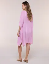 Vestido playero tipo túnica de corte holgado color rosado, confeccionado en tejido semi transparente con textura sutilmente arrugada. Cuenta con escote en V pronunciado, mangas estilo kimono de caída fluida y aberturas laterales en el ruedo irregular.