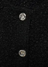 Chaqueta de tejido bouclé en color negro con textura y detalles brillantes. Presenta un diseño recto, cuello redondo, manga larga, cierre frontal con botones efecto joya y dos bolsillos de parche delanteros.