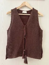 Chaleco tejido en punto calado con hilo de algodón y lurex, de color negro con destellos plateados. Presenta escote profundo en V, cierre frontal con cordones entrelazados terminados en borlas y un corte recto y holgado.