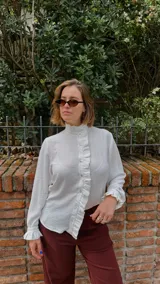 Camisa blanca de textura sutil, con cuello alto tipo victoriano y detalle de volados asimétricos a lo largo de la botonadura frontal. Presenta mangas largas con puños terminados en volados.