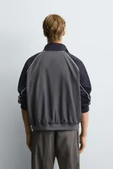 Cazadora relaxed fit en color block, con cuerpo gris oscuro y mangas azul marino. Presenta cuello subido, manga larga, bolsillos frontales de vivo, vivos combinados a contraste en blanco, acabados con rib elástico y cierre frontal con cremallera.