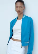Chaqueta tipo bomber color turquesa con cierre frontal de cremallera, cuello redondo y puños con detalles de rayas blancas en contraste. Presenta un diseño recto con dos bolsillos laterales.