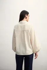 Camisa a rayas verticales color beige y blanco, con cuello clásico, cierre frontal con botón dorado, mangas largas abullonadas con puños elásticos, dos bolsillos frontales y corte holgado.