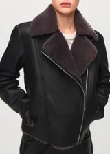 Chaqueta estilo aviador de cuero sintético color negro, con interior y cuello de corderito sintético en tono gris oscuro. Cuenta con cierre frontal cruzado, bolsillos con cierre y detalles de hebillas en las muñecas.