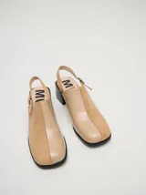 Zuecos de cuero vacuno con acabado en charol, color beige. Presentan un diseño clásico con taco cuadrado de madera de 5 cm y correa trasera ajustable con hebilla metálica.