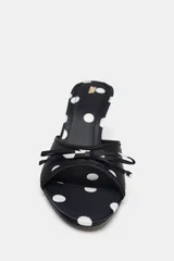 Sandalia tipo mule negra con estampado de lunares blancos, efecto satinado, punta fina, tacón fino y tira delantera con lazo decorativo.