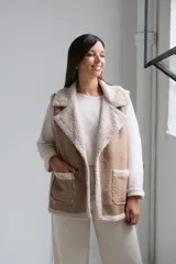 Chaleco de gamuza color beige con interior y detalles de corderito color crudo. Presenta solapas amplias, bolsillos frontales aplicados y diseño sin mangas.