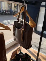 Cartera tipo tote bag de cuero marrón oscuro, con diseño artesanal, costuras visibles en los bordes, bolsillo frontal con flecos y asas trenzadas.