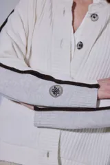 Saco de punto acanalado en color crema, con botones frontales y detalles de ribetes en color marrón oscuro en las mangas. Incluye un parche circular bordado en la manga.