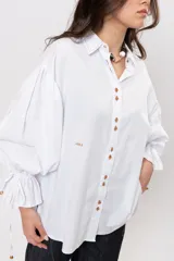 Camisa de algodón blanca de silueta amplia, con cuello camisero, botonadura frontal con botones decorativos, mangas abullonadas extragrandes y detalles de tiritas ajustables con cuentas de resina en los puños.