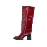 Bota de caña alta Korium modelo Sira, confeccionada en cuero sintético negro con acabado brillante. Presenta un taco cuadrado robusto, punta redondeada y una tira decorativa con hebilla metálica en la zona del tobillo.