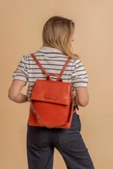 Mochila tipo cartera con tapa, confeccionada en símil cuero color suela. Cuenta con doble compartimento, división interna, cierre de seguridad y bolsillo trasero con cierre. Incluye una correa frontal decorativa con tachas metálicas.