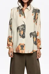 Camisa de corte recto con cuello clásico, cierre frontal de botones y manga larga con puños abotonados. Presenta un estampado integral de perros salchicha sobre fondo color crema.