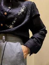 Camisa negra de lino natural, con mangas largas abullonadas, cuello clásico y cierre frontal de botones. Presenta delicados bordados artesanales a mano con motivos florales y detalles brillantes en el frente.