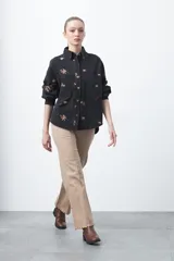 Pantalón de fit recto color beige, confeccionado en cotton spandex. Presenta costuras visibles y bordados decorativos en tono dorado/marrón claro. El ruedo del pantalón tiene un borde festoneado.