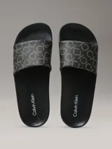 Ojotas Calvin Klein con diseño de monograma de la marca en color beige sobre fondo negro. Presentan una suela anatómica contorneada para mayor confort y una tira superior de material sintético.