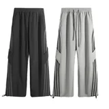 Pantalón deportivo gris claro, estilo cargo o jogging, con franjas negras verticales en los laterales de las piernas. Posee tobillos regulables y cintura ajustable.