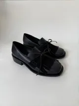 Mocasín de cuero marrón con textura granulada, diseño de horma cuadrada, detalle de flecos y lazo decorativo en el empeine, montado sobre una suela y taco bajo de color negro.