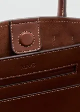 Cartera de mano color marrón, de diseño estructurado tipo trapecio, con doble asa corta y detalles de correas laterales ajustables con hebilla.