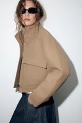 Cazadora corta de paño color tostado, con cuello alto, mangas largas, bolsillos frontales con solapa y cierre con cremallera oculta y botones a presión metálicos.